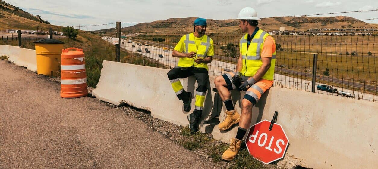 Zwei Arbeiter sitzen auf einer Betonabsperrung am Stra&szlig;enrand. Sie tragen kurze Warnschutzkleidung und K&uuml;hlwesten. Im Hintergrund sieht man eine karge Berglandschaft und bew&ouml;lkten, blauen Himmel. 