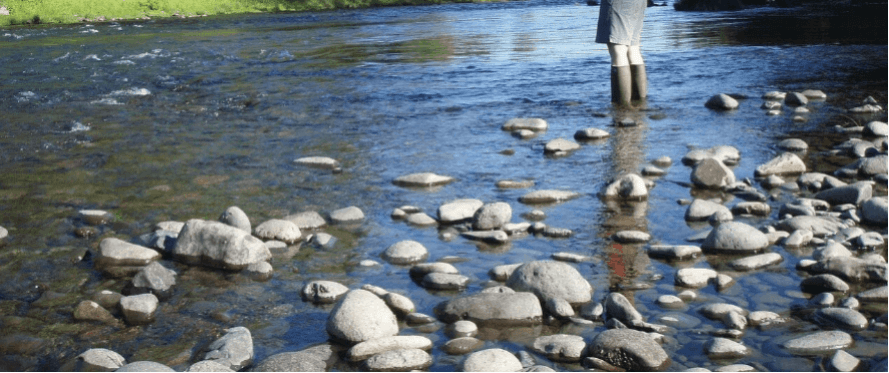 Man sieht ein Flussbett mit großen Steinen und klarem, fließendem Wasser. Im rechten hinteren Bildbereich steht eine Person in kurzer Hose und grünen Gummistiefeln.