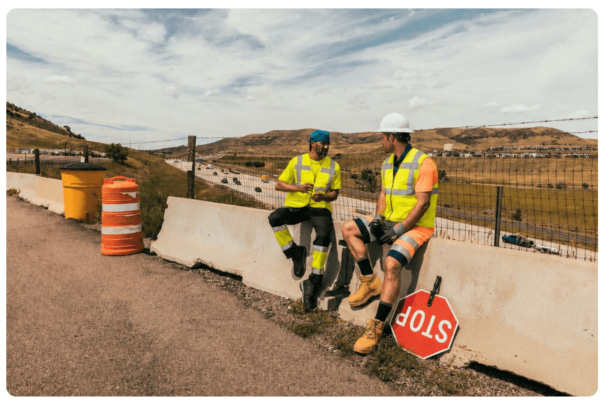Zwei Arbeiter sitzen auf einer Betonabsperrung am Straßenrand. Sie tragen kurze Warnschutzkleidung und Kühlwesten. Im Hintergrund sieht man eine karge Berglandschaft und bewölkten, blauen Himmel. Ein Link zu unseren Kühlwesten ist hinterlegt.