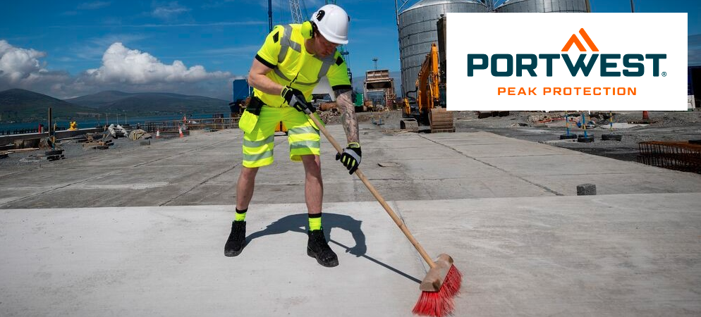 Mann in warngelber Arbeitskleidung und wei&szlig;em Schutzhelm, der vor einer Fabrik den Betonboden kehrt. Im Hintergrund sieht man Berge und blauen Himmel mit kleinen Wolken. Im rechten oberen Eck ist das blau-orange Logo des Herstellers Portwest platziert.