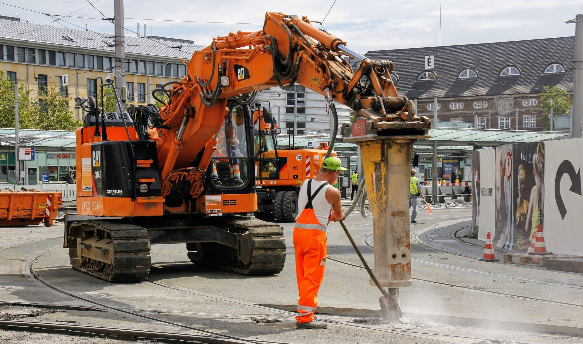 Urbane Baustelle mit H&auml;usern und Busbahnhof im Hintergrund. Zentral stehen ein oranger Bagger und ein Arbeiter in Warnschutzkleidung. 