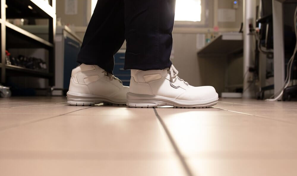 Man sieht ein Paar Beine in schwarzer Hose mit wei&szlig;en Schuhen. Im Hintergrund sieht man einen Fliesenboden und K&uuml;chenequipment aus Edelstahl.