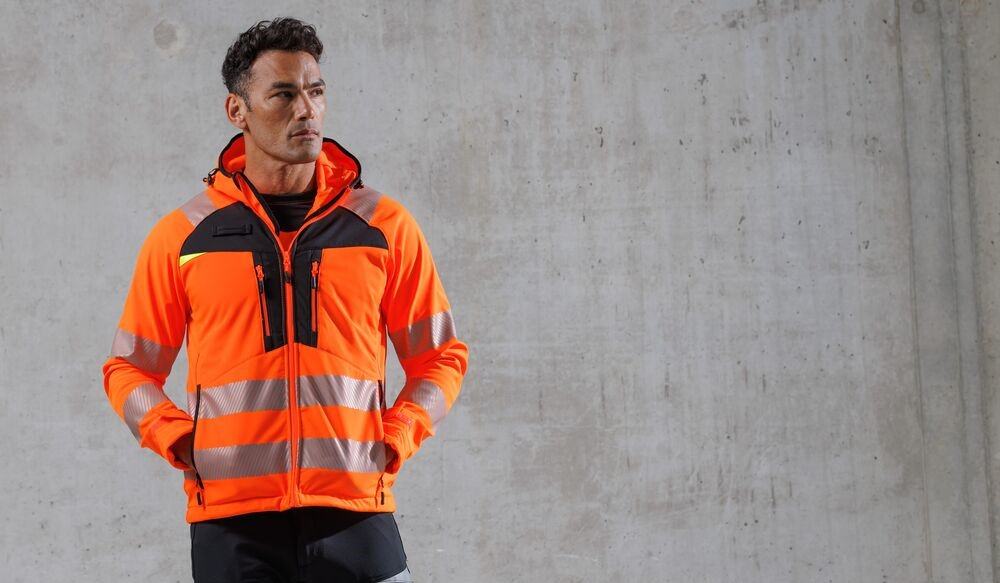 Mann in warnoranger Jacke. Er hat die Hände in den Taschen und blickt nicht direkt in die Kamera. Im Hintergrund sieht man eine graue Wand.