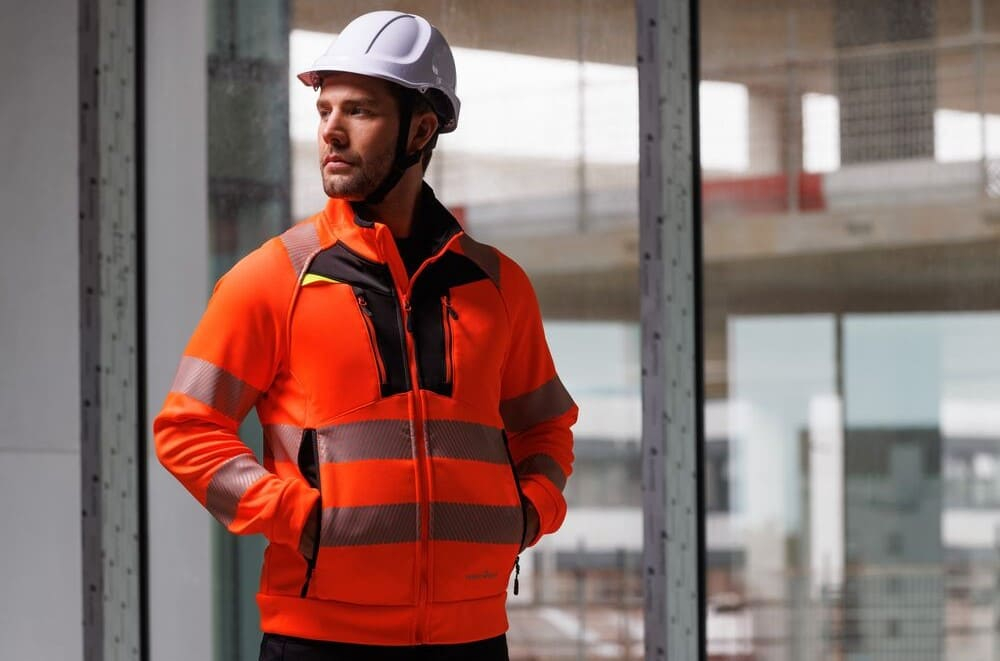 Ein Mann in orangem Warnschutz-Sweatshirt und weißem Schutzhelm steht vor einem Fenster. Draußen sieht man ein Haus im Rohbau.