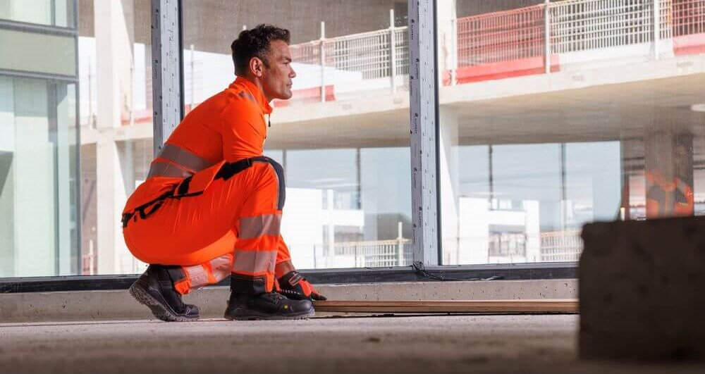 Ein Mann in oranger Warnschutzkleidung und schwarzen Sicherheitsstiefeln hebt aus der Hocke etwas vom Boden auf. Im Hintergrund sieht man ein Haus im Rohbau.