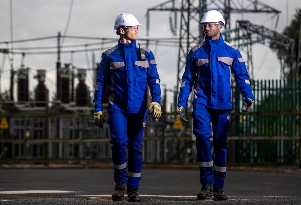 Zwei Arbeiter in blauer flammhemmender Kleidung und wei&szlig;en Schutzhelmen. Sie unterhalten sich und laufen auf die Kamera zu. Im hintergrund sieht man ein E-Werk.