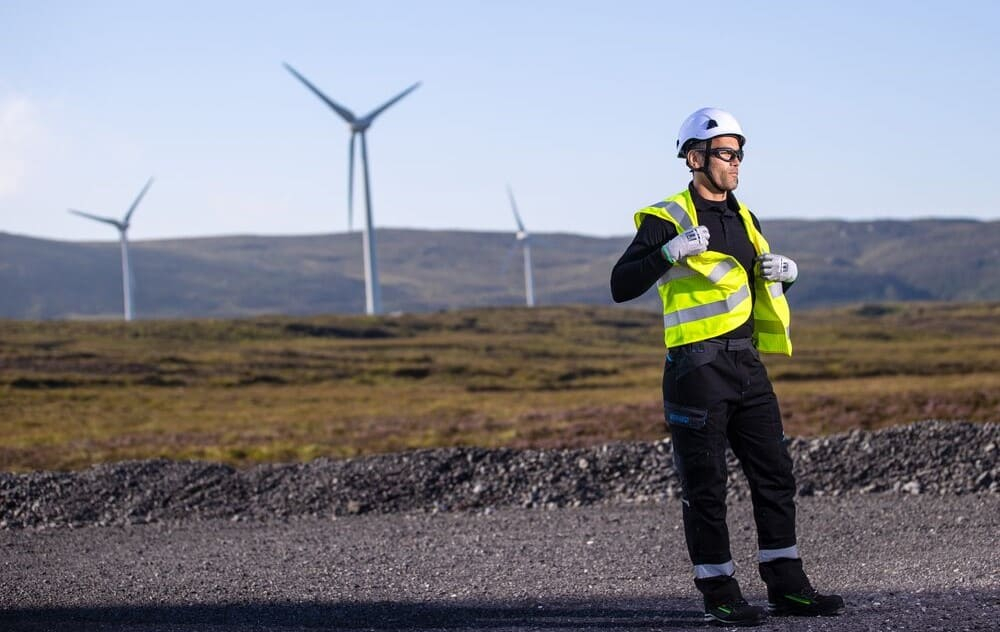 Ein Mann in schwarzer Arbeitskleidung, wei&szlig;em Schutzhelm und gelber Warnweste steht vor einem Windpark.