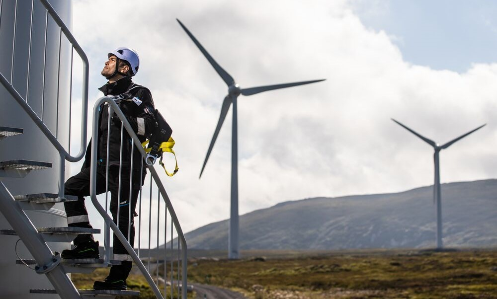 Ein Arbeiter in schwarzer Arbeitskleidung steigt die Stufen zu einem Windrad hinauf. Er trägt einen weißen Schutzhelm und ist mit Fallsicherung ausgestattet. Im Hintergrund sieht man Berge und zwei Windräder.