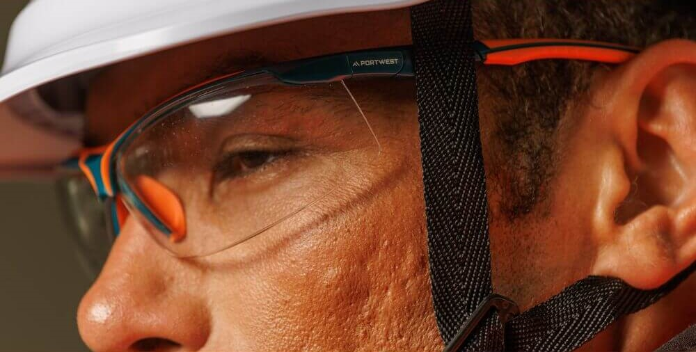 Nahaufnahme eines Mannes, der einen weißen Schutzhelm und eine klare Brille mit orangen und schwarzen Details trägt.