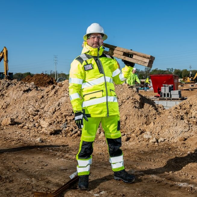 Mann in warngelber Arbeitskleidung der auf einer Baustelle steht. Der Himmel über ihm ist blau.