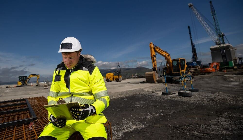 Mann in warngelber Arbeitskleidung, weißem Schutzhelm und Anti-Impact-Handschuhen sitzt auf einem Stahlgitter vor einer Baustelle und schaut sich etwas auf einem Klemmbrett an. Im Hintergrund sieht man Bagger, Kräne und andere Baumaschinen.