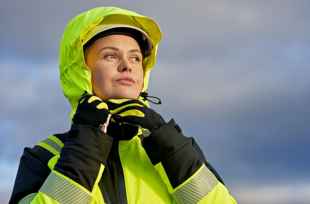 Eine Arbeiterin in warngelber Schutzkleidung, Handschuhen, Helm und Kapuze. Sie hat beide Hände am Kinn. Der Himmel im Hintergrund ist blau und wolkig.