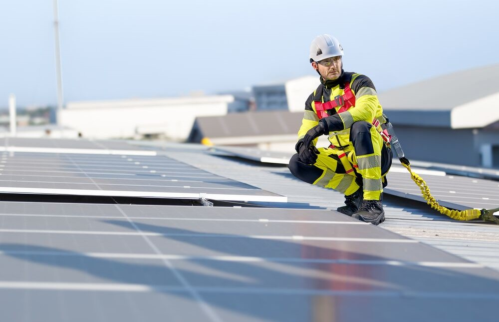 Ein Arbeiter in gelber Warnschutzkleidung und weißem Schutzhelm kniet vor einer Fotovoltaikanlage. Im Hintergrund sieht man einen blassblauen, wolkenlosen Himmel.