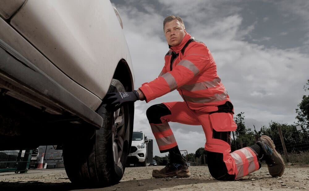 Ein Mann in oranger Warnschutzkleidung kniet vor einem Auto und prüft einen Reifen. Im Hintergrund sieht man andere Autos und blauen, leicht bewölkten Himmel.