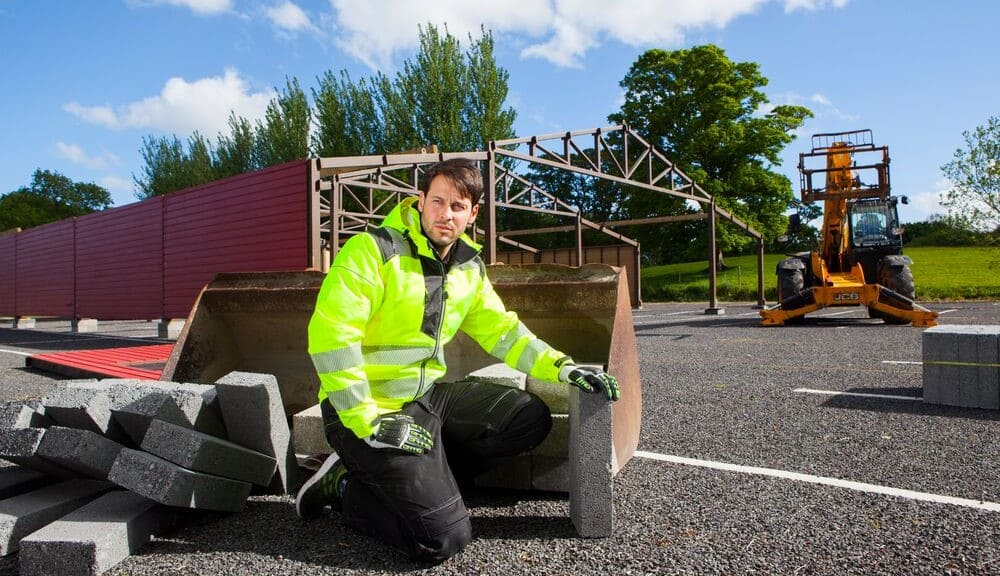 Mann in gelber Warnschutzjacke kniet auf einer Baustelle auf dem Boden. Hinter ihm sieht man eine halbfertige Halle und eine Baumaschine.