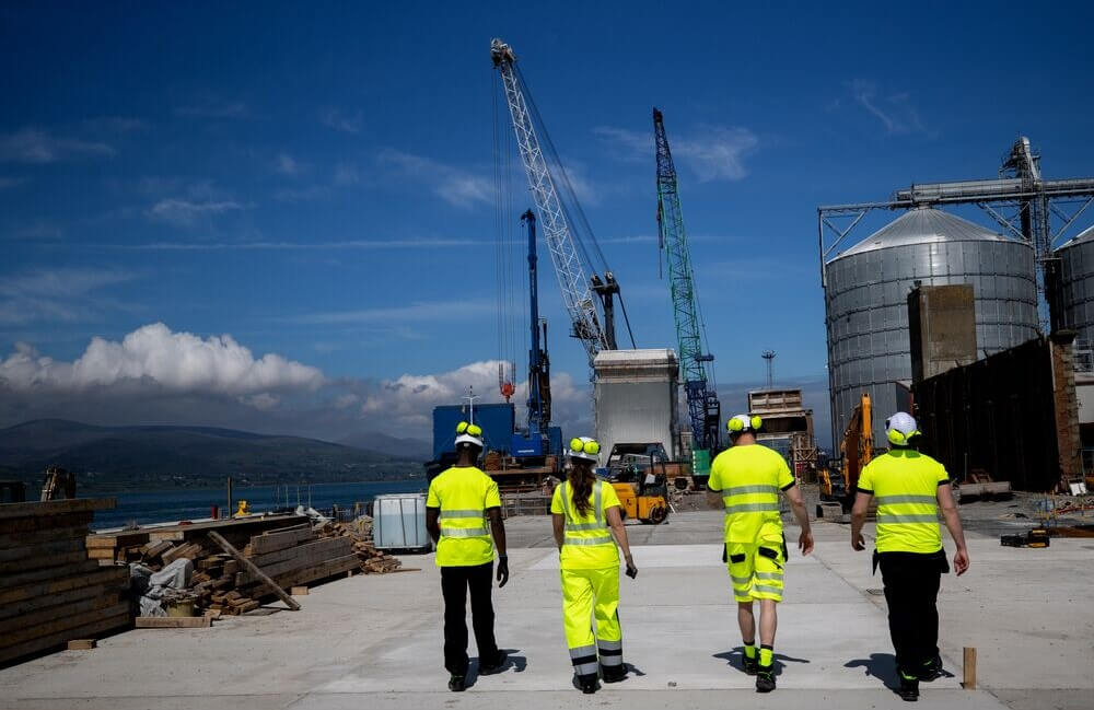 R&uuml;ckenansicht vierer Arbeiter in warngelber Arbeitskleidung. Im Hintergrund sind Kr&auml;ne, ein Silo, Baumaschinen und eine Baustelle sowie eine Bergkette und blauer Himmel mit kleinen Wolken zu sehen.