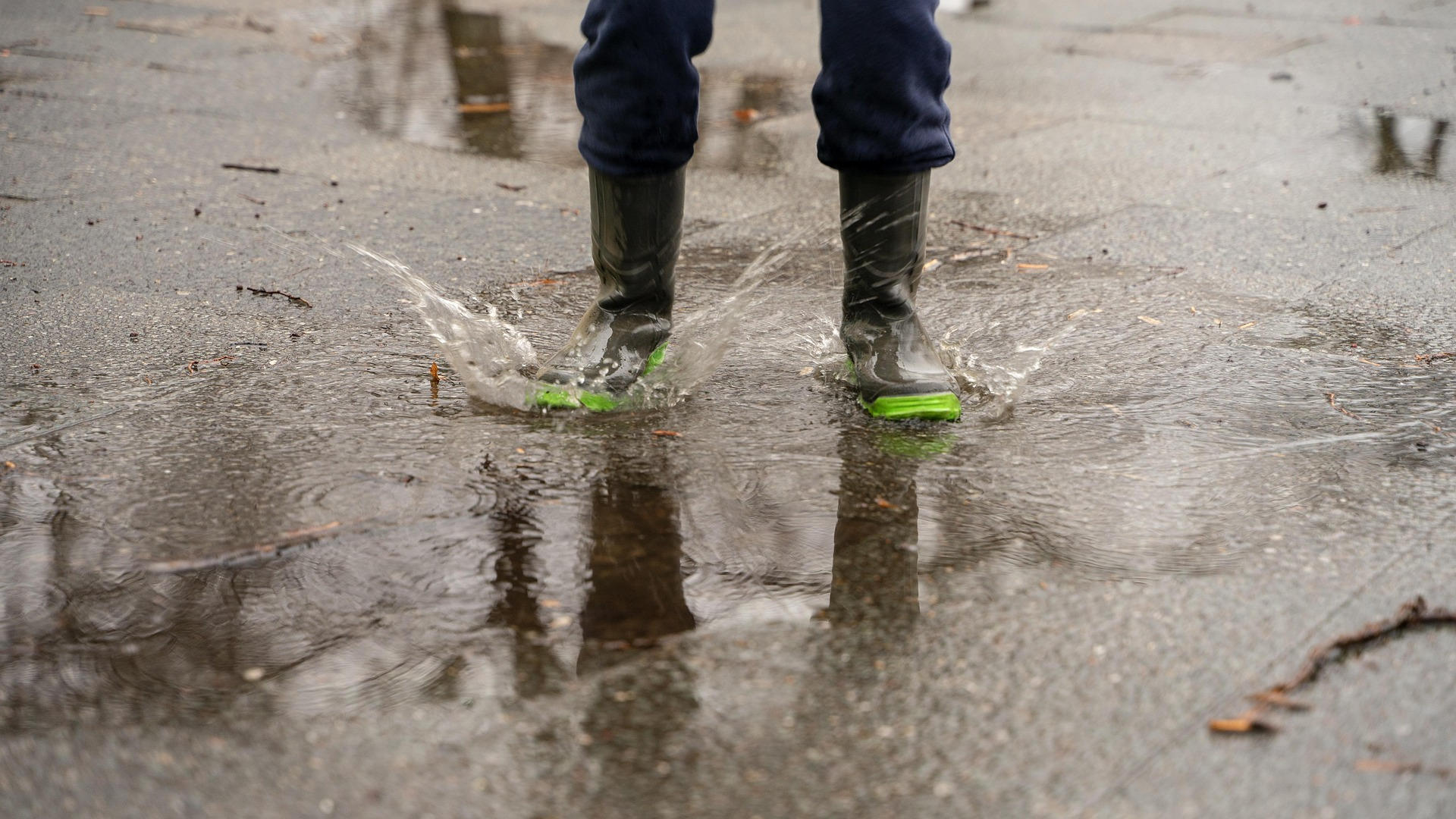 Man sieht eine Person in grünen Gummistiefeln in einer Pfütze auf nassem Asphalt.