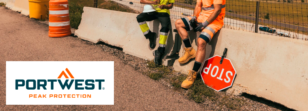 Man sieht die Beine zweier Personen in Arbeitskleidung. Sie sitzen auf einer Betonabsperrung, ein Stopschild liegt neben Ihnen und im Hintergrund befindet sich ein Zaun, eine Straße und eine Hügelkette. Das Logo des Herstellers Portwest in ORange und Blau auf weißem Grund befindet sich im linken unteren Bildrand. 