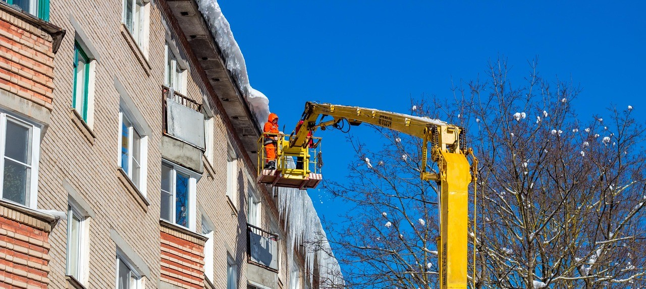 Man sieht eine Häuserreihe mit Eiszapfen am Dachfirst und Schnee auf dem Dach. Auf einer Hebebühne steht ein Arbeiter in oranger Wanrschutzkleidung. Rechts sieht man einen kahlen Baum mit Schnee auf den Ästen.