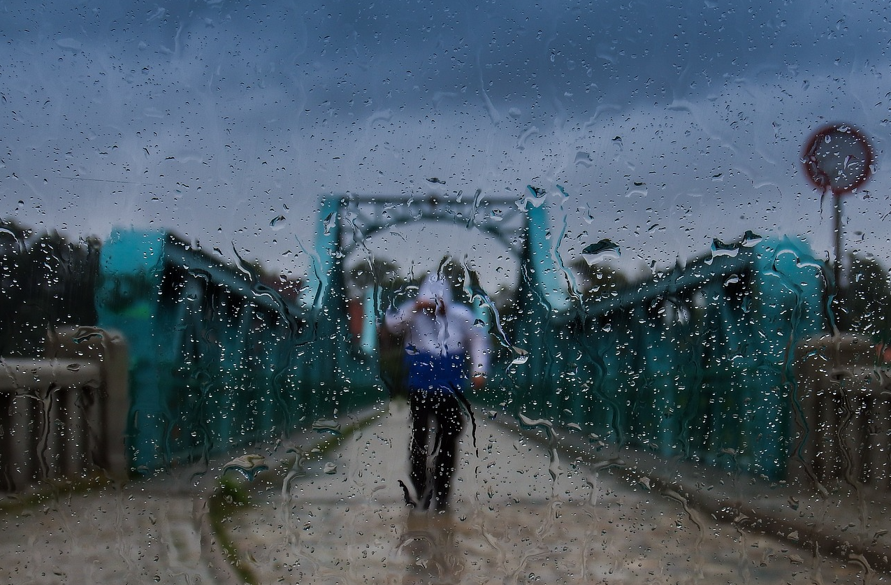 Blaue Brücke vor grauem Himmel mit einer Person in Regenbekleidung in der Mitte der Straße. Die Szene wurde durch eine nasse Glasscheibe fotografiert. Ein Link zu unserer gesamten Auswahl praktischer Regenbekleidung ist hinterlegt.