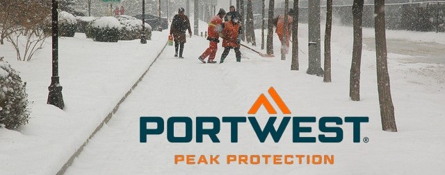 Schneebedeckter Gehweg mit dick eingepackten Fußgänger:innen, Bäumen und Menschen in Warnkleidung, die Schnee schaufeln.