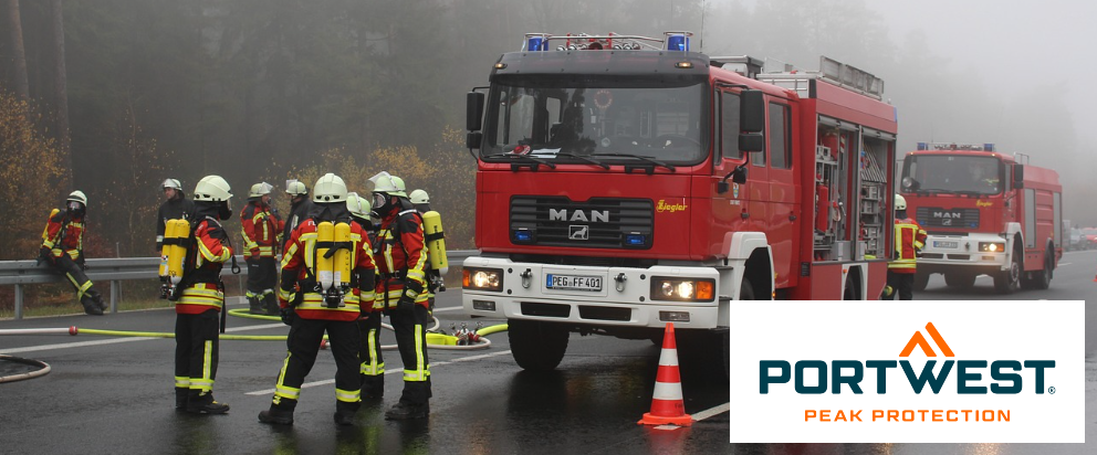 Straßenszene mit zwei roten Feuerwehrautos und diversen Feuerwehrleuten. Im Hintergrund sieht man nebelverhangene Bäume. Im rechten unteren Eck befindet sich das blau-orange Logo der Firma Portwest vor einem weißen Rechteck.