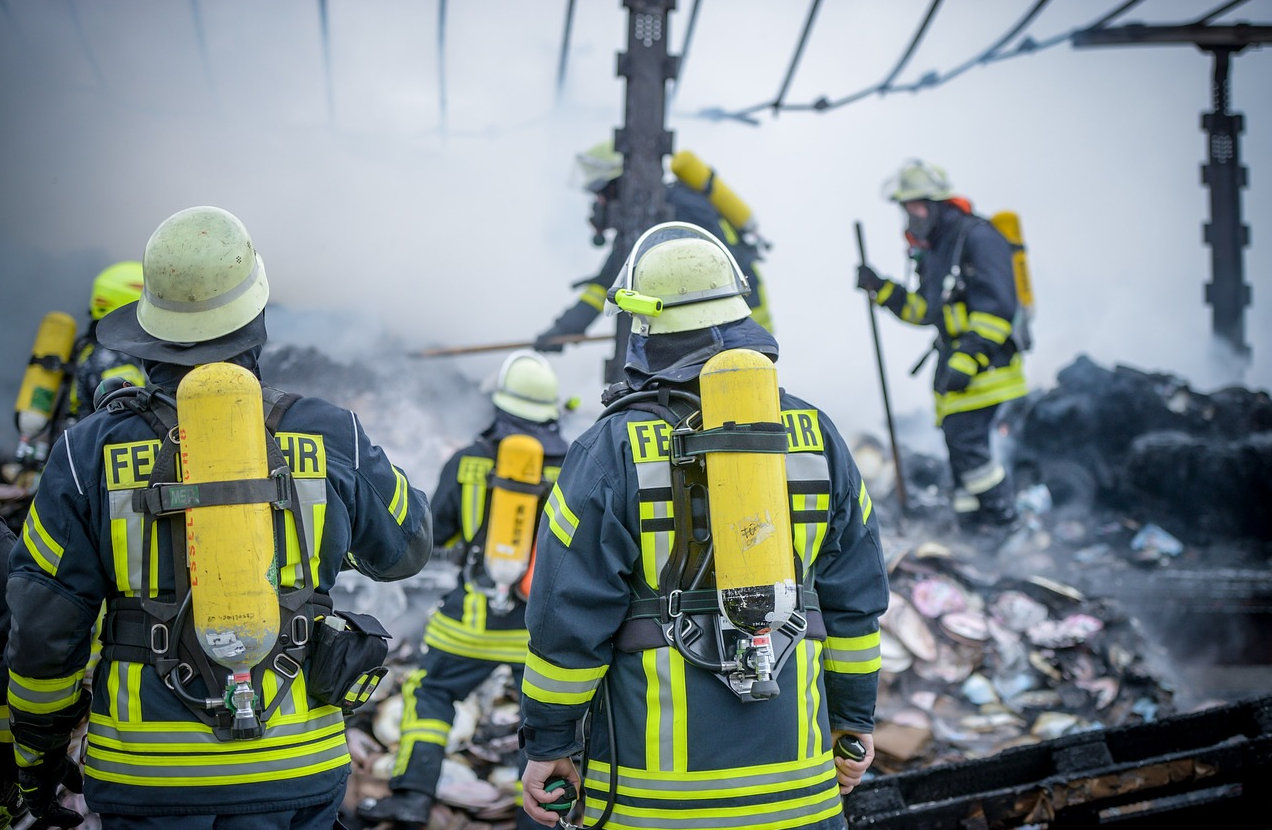 Feuerwehrleute in blauer Kleidung mit gelben Warnstreifen und Helmen auf einem Schrottplatz. Der hinterlegte Link führt zu unserer Auswahl an flammhemmender Feuerwehrkleidung. 