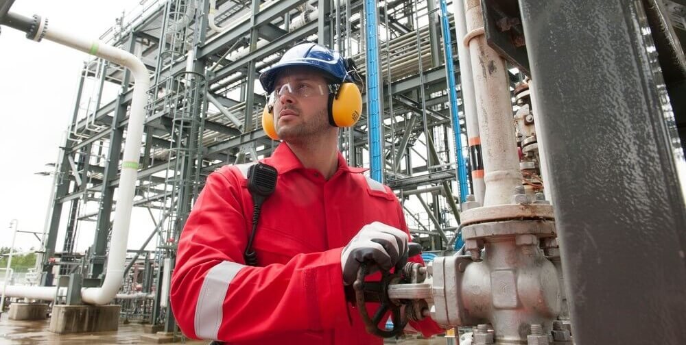 Ein Arbeiter in roter Warnschutzkleidung, blauem Schutzhelm und gelbem Geh&ouml;rschutz dreht an einem Ventilrad. Im Hintergrund sieht man Rohre und eine Stahlkonstruktion.