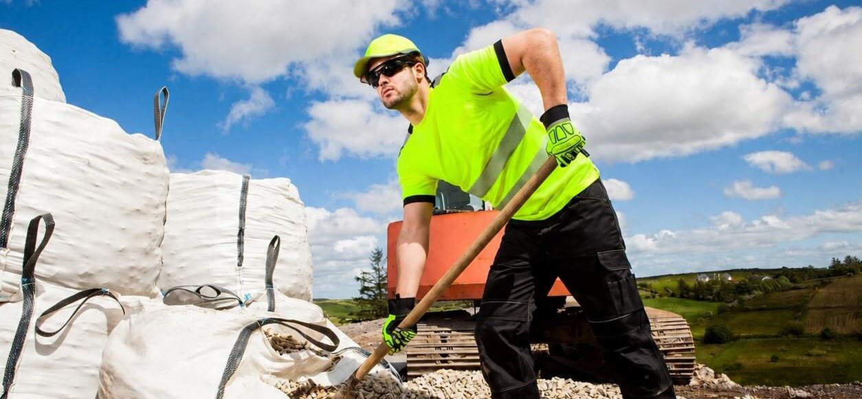 Mann in gelbem Shirt und schwarzer Hose. Er hat eine gelbe Kappe auf dem Kopf, trägt eine Sonnenbrille und schaufelt Kies aus großen Säcken. Im Hintergrund sieht man sanft bewölkten, blauen Himmel eine Baustelle und grüne Hügel.