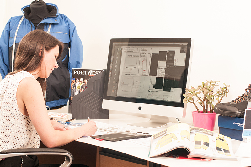 Brünette Frau bei der Arbeit am Schreibtisch. Auf ihrem Computerbildschirm finden sich technische Zeichnungen von Arbeitskleidung.