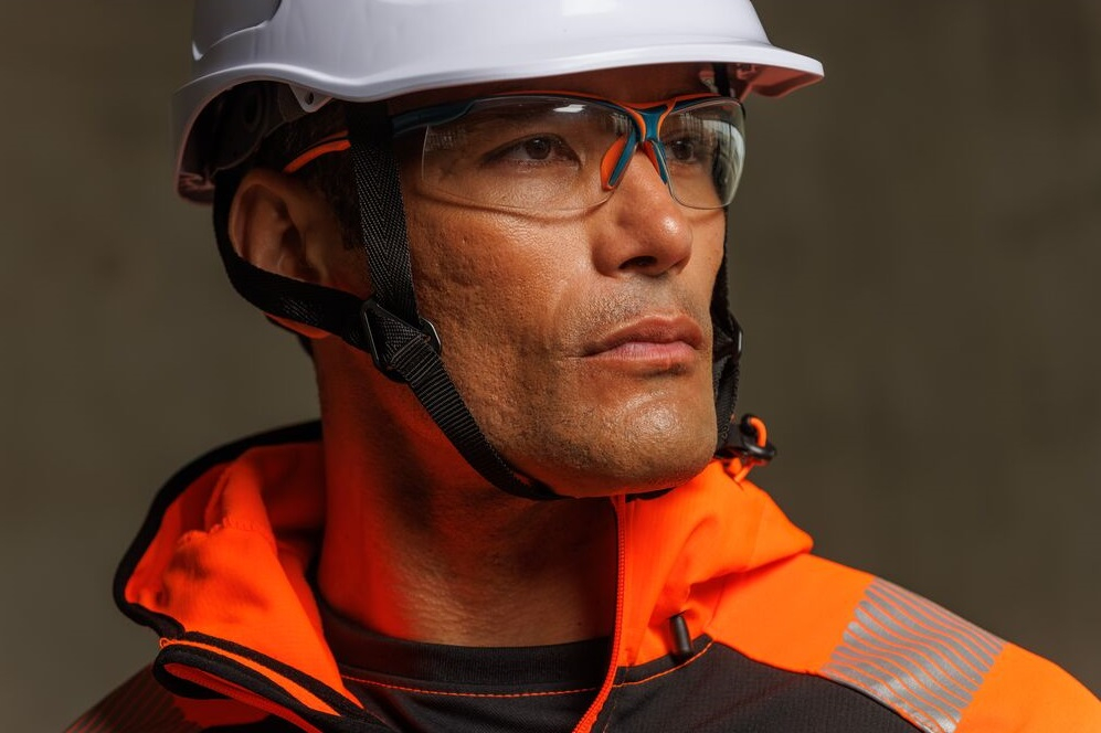 Nahaufnahme eines Arbeiters mit weißem Schutzhelm und klarer Schutzbrille, der konzentriert in die Ferne blickt; trägt leuchtend orange-schwarze Warnschutzkleidung. Ein Link zu unserer Kollektion an Augenschutz ist hinterlegt.