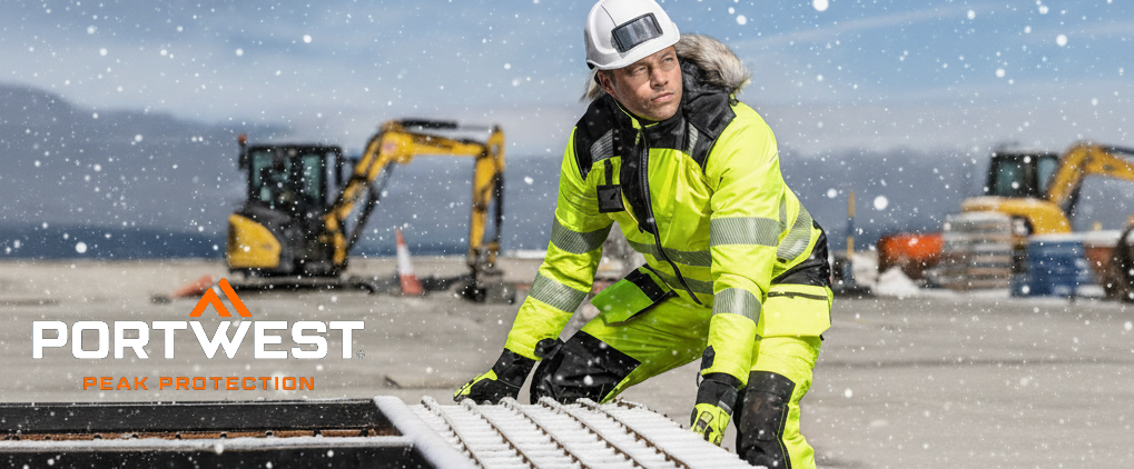 Arbeiter in gelber, reflektierender Winter-Arbeitsschutzkleidung arbeitet im Freien bei Schneefall. Er tr&auml;gt Schutzhelm und Handschuhe. Im Hintergrund sind Baustellenfahrzeuge und verschneite Landschaft zu sehen. Im linken unteren Eck befindet sich das Logo des Herstellers Portwest in wei&szlig;er und oranger Schrift.