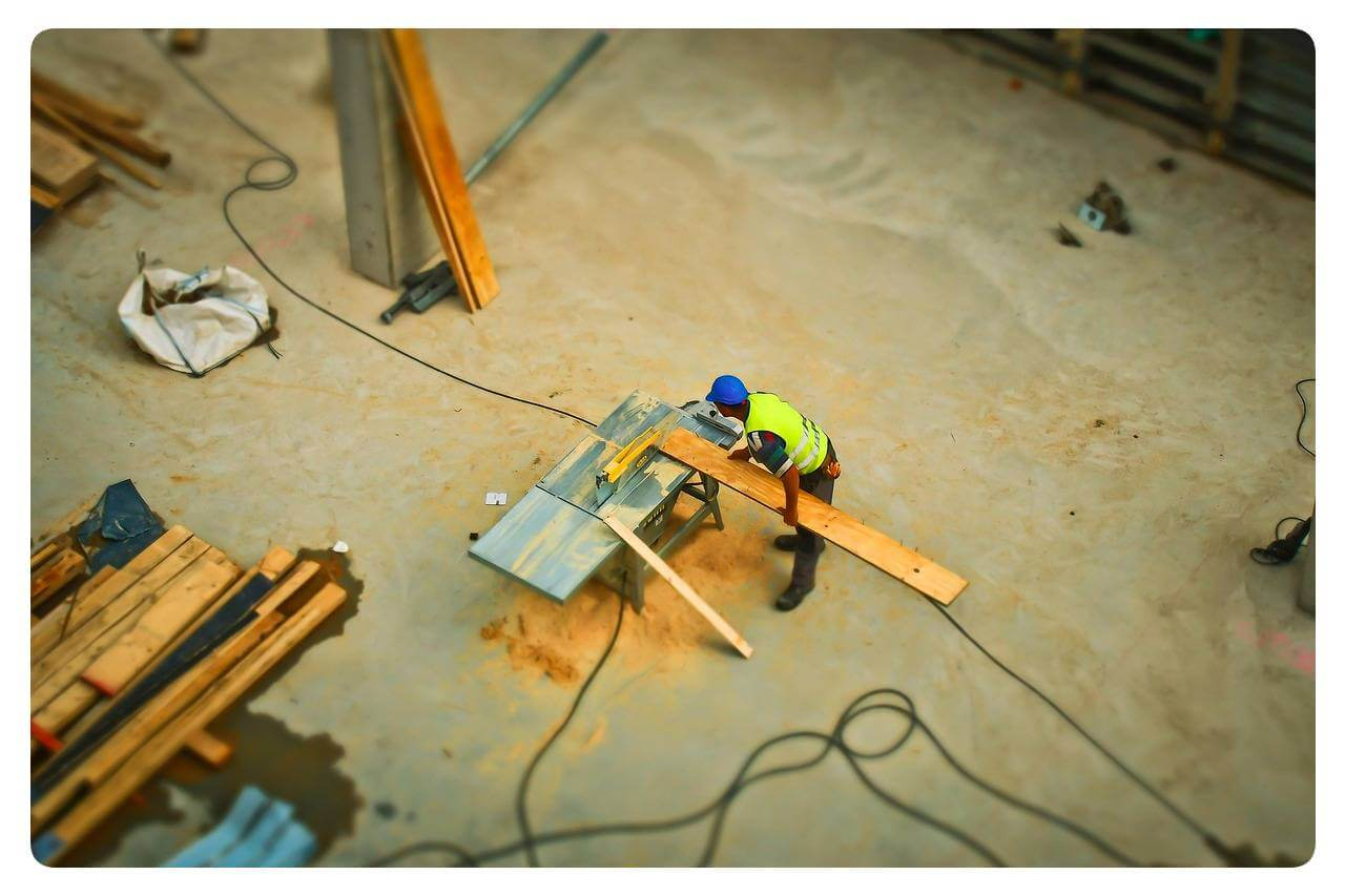 Man sieht einen Arbeiter in gelber Warnweste aus der Vogelperspektive. Er s&auml;gt ein Holzbrett an einer Tischkreiss&auml;ge.