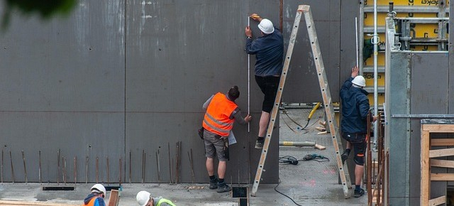 Man sieht zwei Arbeiter auf einer Baustelle. Einer steht auf einer Leiter, der andere hält die Leiter fest. Beide tragen kurze Arbeitshosen, einer trägt einen weißen Helm und der andere eine orange Warnschutzweste.