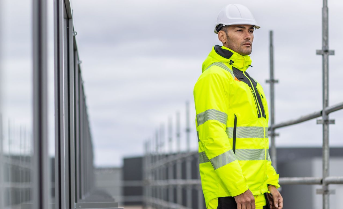 Arbeiter in warngelber Arbeitskleidung mit weißem Helm vor einer Fensterfront und dem oberen Teil eines Gerüstes mit hinterlegtem Link zur DX4-Kollektion.