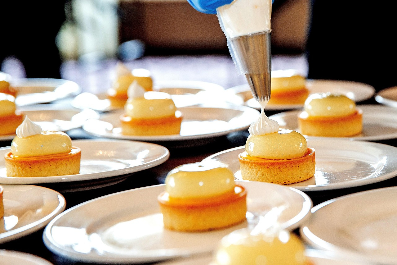 Teller mit feinem Gebäck. Eine Hand in blauem Handschuh hält einen Spritzbeutel und spritzt eine weiße Masse auf ein Törtchen.