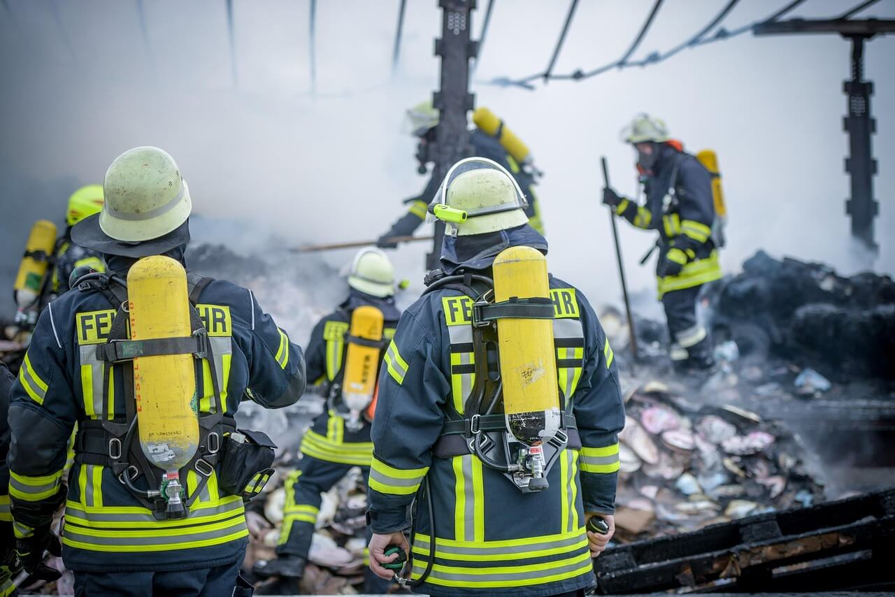 Sechs Feuerwehrleute in voller Montur bei einem Einsatz. Man sieht verkohlte Tr&uuml;mmer und undefinierbare Eisengestelle.Ein Link zu unserer Feuerwehrkleidung ist hinterlegt.