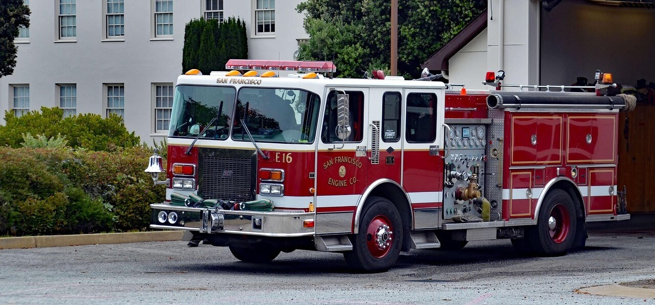 Man sieht ein Feuerwehrauto aus San Francisco, das aus einer Garage fährt. Im Hintergrund ist ein Haus, das von grünen Büschen gesäumt wird.