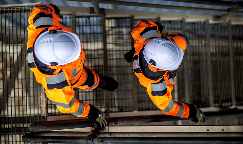 Vogelperspektive zweier Arbeiter mit weißen Schutzhelmen, schwarz-grünen Handschuhen, Sicherheitsstiefeln und Warnschutzoverall in Orange mit Link zu unserer Kollektion Störlichtbogen-Schutzkleidung. 