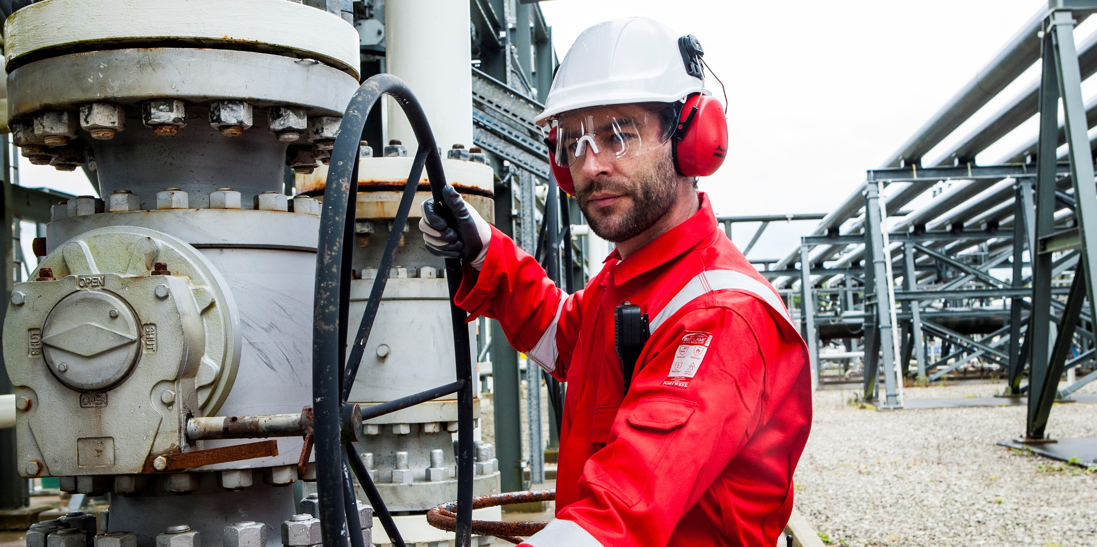 Arbeiter in industriellem Kontext in Schutzkleidung: weißer Helm, Brille, roter Ohrenschutz, schwarze Handschuhe und Schutzoverall FR50 in Rot mit Reflexstreifen.