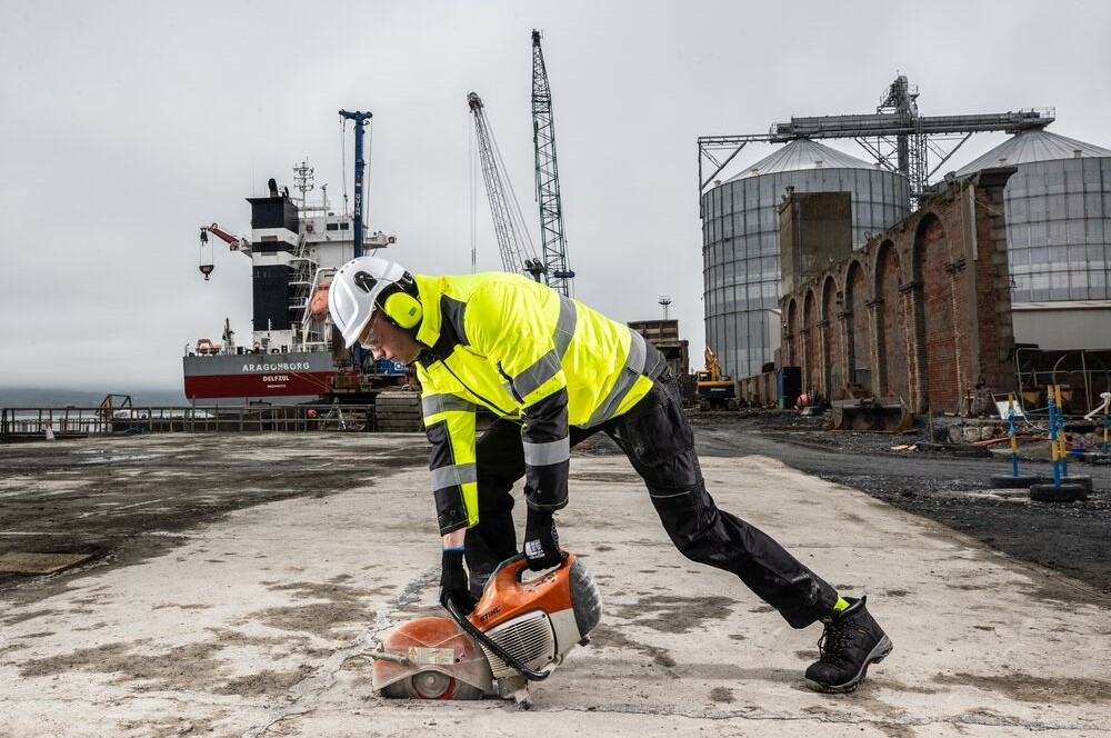 Ein Arbeiter in gelber Warnschutzjacke und schwarzer Arbeitshose, der eine Bodenplatte mit einer Kreiss&auml;ge s&auml;gt. Im Hintergrund sieht man gro&szlig;e Silos und ein Containerschiff. Das Wetter ist grau und regnerisch. ein Link zur PW3 Warnschutz 3-in-1 Jacke PW365 ist hinterlegt.