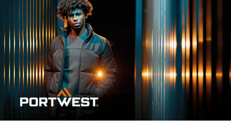 Ein Arbeiter in schwarzer Arbeitskleidung vor abstraktem Hintergrund mit gestreifter Wand und Lichtreflexen. Das Logo des Herstellers Portwest in Wei&szlig; und Orange befindet sich im linken unteren Bildrand.