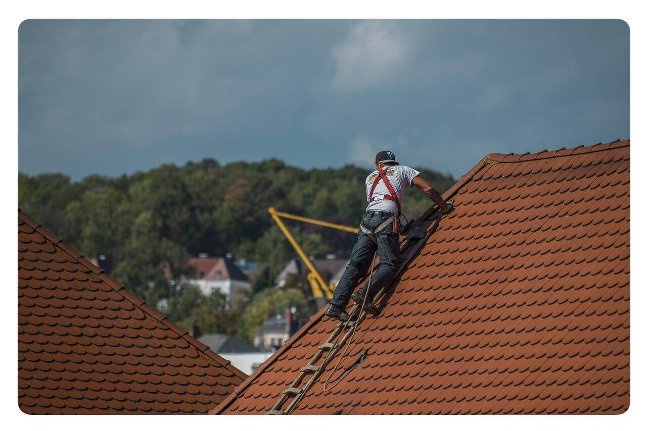 Man sieht einen Arbeiter, der auf einer Leiter auf einem roten Dach steht. Er ist mit einer Absturzsicherung gesichert.