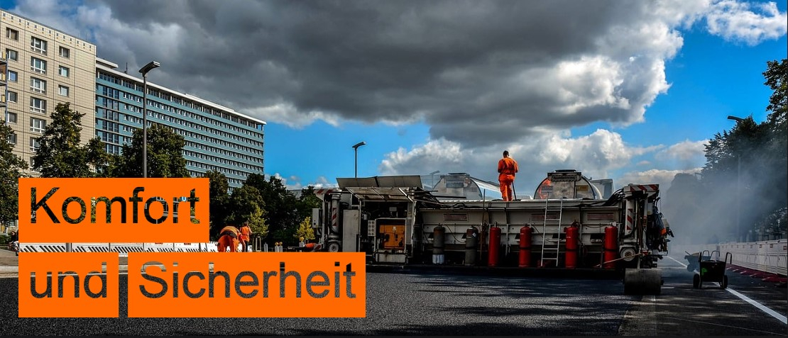 Szene einer Baustelle im Straßenbau. Links im Bild sieht man Hochhäuser und einige Bäume, der rechte Bildbereich ist von einer Wasserfontäne bedeckt. Im linken unteren Bildrand stehen die Worte "Komfort und Sicherheit" in orange hinterlegten, ausgeschnittenen Buchstaben. 