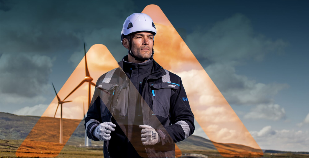 Mann mit Schutzhelm, Handschuhen und Arbeitsjacke steht vor Windkraftanlagen; Darstellung von Arbeitssicherheit und Absturzsicherung bei T&auml;tigkeiten in der H&ouml;he.