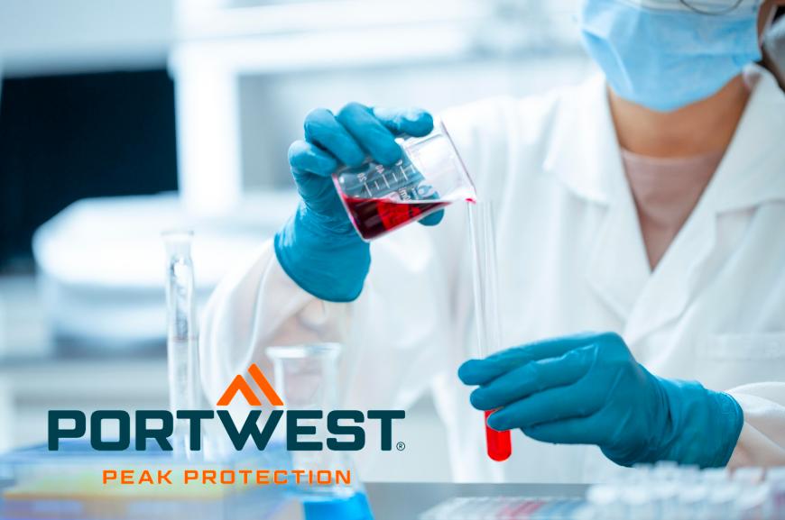 Labormitarbeiterin in Schutzkleidung mit Laborkittel, blauen Handschuhen und Mundschutz füllt rote Flüssigkeit aus einem Becherglas in ein Reagenzglas. Portwest-Logo mit Slogan ‚Peak Protection‘ im Vordergrund. Ein Link zu unserer Auswahl an Laborkitteln ist hinterlegt.
