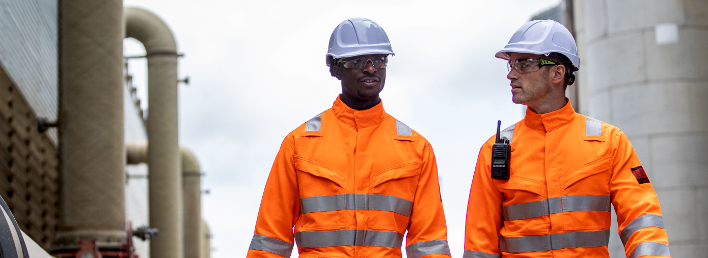 Zwei Arbeiter vor industriellem Hintergrund, die Warnschutzkleidung in Orange und weiße Schutzhelme tragen. 