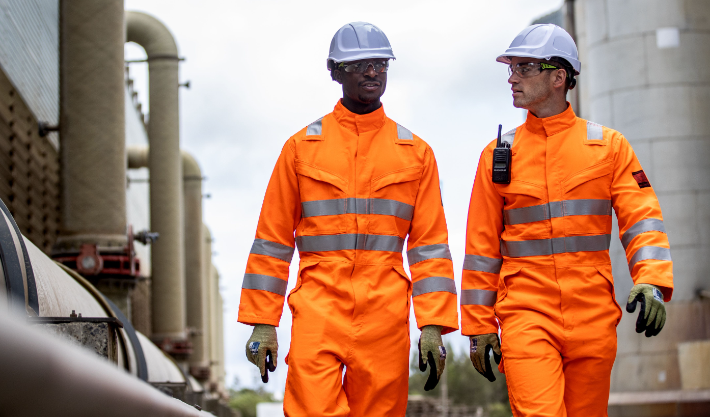 Zwei Arbeiter vor industriellem Hintergrund, die Warnschutzkleidung in Orange und weiße Schutzhelme tragen. Ein Link zum Endurance Glowtex Helm PG54 ist hinterlegt.