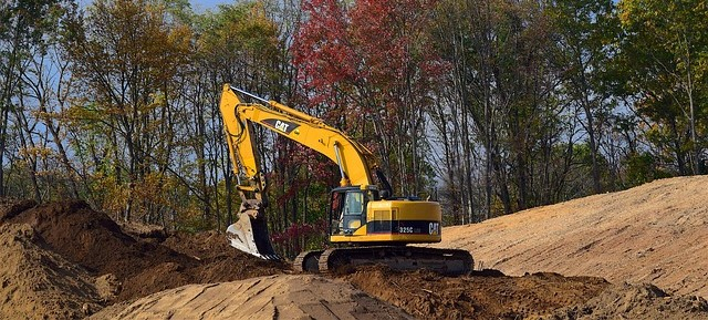 Man sieht einen gelben Bagger inmitten einer sandigen Baustelle. Im Hintergrund sieht man B&auml;ume mit gr&uuml;nem und buntem Laub und blauen Himmel.