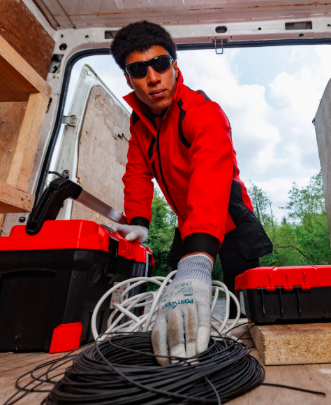 Arbeiter in Sonnenbrille, roter Softsheljacke, schwarzer Cargo-Hose und weißen Handschuhen streckt sich in den Innenraum eines weißen Transporters, um ein zusammengerolltes Bündel schwarzer Kabel zu greifen. Im Hintergrund sieht man blauen Himmel mit Wolken und frisch grüne Bäume. Hinterlegt ist ein Link, der zu unserer Auswahl a ESD-Handschuhen führt.